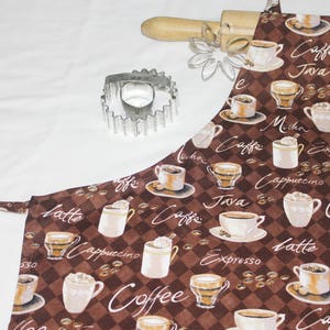 May include: A brown and white kitchen apron with a checkered pattern and coffee cup designs. The apron has the words "Caffe", "Java", "Cappuccino", "Latte", "Espresso", and "Coffee" printed on it.
