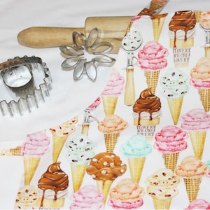 May include: White cotton apron with a repeating pattern of colorful ice cream cones. The text "I love ice cream" is printed on the cones. The apron is shown with a rolling pin, a flower-shaped cookie cutter, and a round cookie cutter.