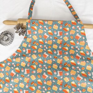 May include: A teal blue apron with a pattern of colorful cakes, cupcakes, and donuts. The apron has adjustable straps and a large pocket in the front.