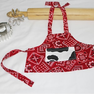 May include: Red bandana print children's apron with a white cow print pocket. The apron has adjustable straps and ties at the waist.