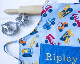 May include: A blue and white construction vehicle print apron with a blue pocket that says "Ripley" in gold embroidery. The apron has adjustable straps and a neck strap.