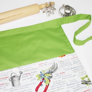 May include: A green apron with a white fabric pocket featuring a print of gardening tools and text about different types of plants. The text includes "Harvest", "All Berry", "Kale", and "Blueberry".