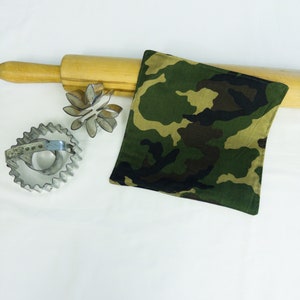 May include: A camouflage fabric kitchen towel or dishcloth. The towel is lying on top of a wooden rolling pin and two metal cookie cutters.