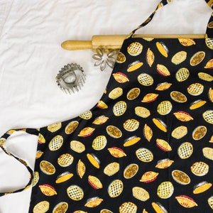 May include: A black apron with a pattern of yellow and red pies. The apron has adjustable straps and a pocket.