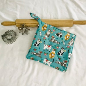 May include: A teal blue fabric pot holder with a repeating pattern of cats and flowers. The cats are in various poses and colors, including white, gray, orange, and black.