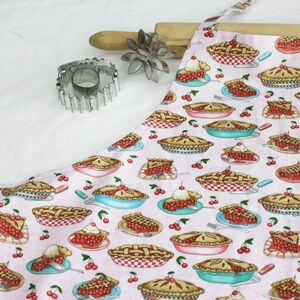 May include: A pink apron with a pattern of cherry pies, slices, and cherries. A rolling pin, a metal pie crust cutter, and a flower-shaped cookie cutter are on the white surface. The apron has ties and a neck strap.
