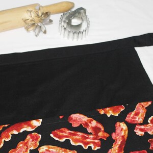 May include: Black apron with a large pocket and a bacon print. The apron has a black fabric top and a bottom section with a red and white bacon print.
