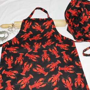 May include: A black apron with a red lobster print and a matching black chef's hat. The apron has adjustable straps and a tie at the waist.