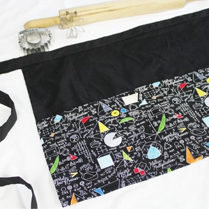 May include: Black apron with a large pocket and adjustable ties. The pocket has a colorful print of mathematical equations, geometric shapes, and symbols.