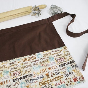May include: Brown apron with a large pocket and adjustable neck strap. The pocket has a white fabric lining with a coffee themed print featuring words like "latte", "mocha", "cappuccino", and "espresso".