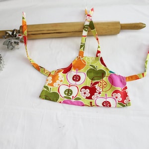 May include: A colorful child's apron with a green background and a pattern of red, orange, and yellow apples. The apron has two long ties and is lying on a white surface next to a wooden rolling pin and two metal cookie cutters.