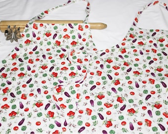May include: Two white aprons with a colorful vegetable print. The print features red tomatoes, green peppers, and purple eggplants on a white background.