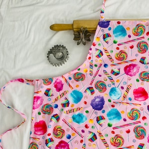 May include: Pink cotton apron with a colorful candy and cotton candy print. The apron has adjustable straps and a large pocket.