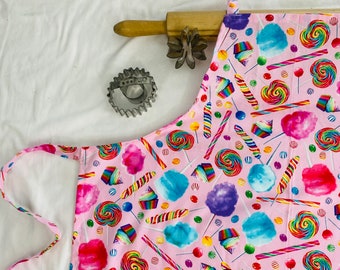 Puede incluir: Delantal de algodón rosa con un estampado colorido de caramelos y algodón de azúcar. El delantal tiene tirantes ajustables y un bolsillo grande.