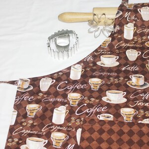 May include: A brown and white kitchen apron with a checkered pattern and a coffee theme. The apron features various coffee cups, coffee beans, and the words "Coffee", "Java", "Espresso", "Cappuccino", "Latte", and "Mocha".