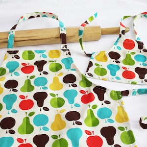 May include: A set of two aprons with a pear and apple pattern. The larger apron is white with brown, green, yellow, and blue pears and apples. The smaller apron is white with red, green, yellow, and blue pears and apples.