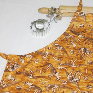 May include: A brown and yellow apron with a dinosaur skeleton pattern. The apron has a long neck strap and a short waist strap.