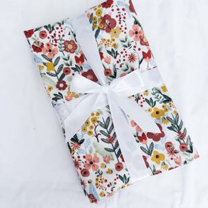 May include: A rectangular gift wrapped in floral patterned paper with a white ribbon tied in a bow. The floral pattern includes red, yellow, and pink flowers with green leaves on a white background. The gift is on a white surface.