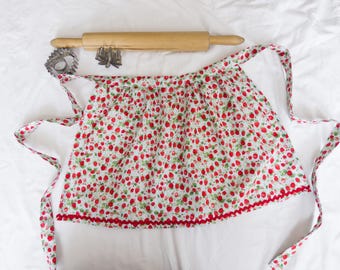 May include: A white apron with a red trim and a strawberry print. The apron has two long ties and a gathered waist.