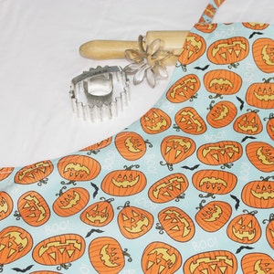 May include: A blue and orange Halloween apron with a pattern of jack-o'-lantern faces. The apron has a tie at the neck and a tie at the waist.