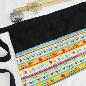 May include: Black apron with a colorful school-themed print featuring books, pencils, apples, and handprints. The apron has a long black strap and multiple pockets.