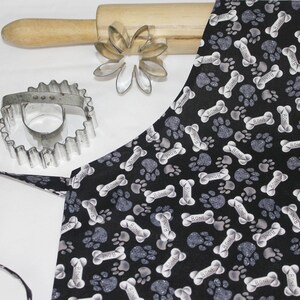 May include: Black and white kitchen apron with a paw print and bone pattern. The apron has a long tie and a short neck strap.
