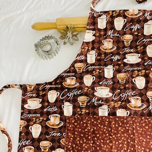 May include: A brown and white kitchen apron with a checkered pattern and a pocket. The apron is decorated with a variety of coffee cups and coffee-related text, including "Mocha", "Java", "Cappuccino", "Latte", "Espresso", and "Coffee".