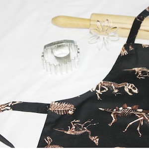 May include: Black apron with a dinosaur skeleton print. The apron has adjustable straps and a large pocket.