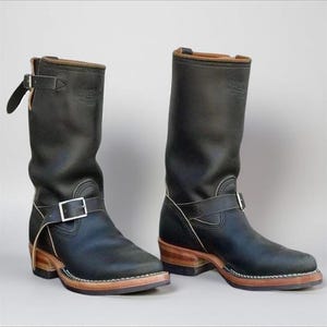 Puede incluir: Un par de botas de moto de cuero negro. Las botas tienen una altura media de pantorrilla, correas con hebilla y una suela resistente con un tacón marrón claro. El cuero tiene un aspecto ligeramente desgastado.