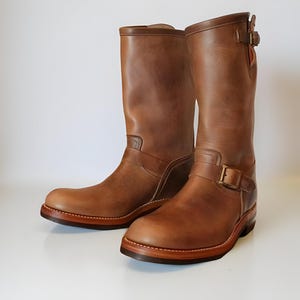 Puede incluir: Un par de botas de cuero marrón con un diseño clásico. Las botas tienen una altura media de pantorrilla, una puntera redondeada y una suela resistente. Cuentan con cierres de hebilla y costuras decorativas. Las botas miden unos 30 cm de alto.