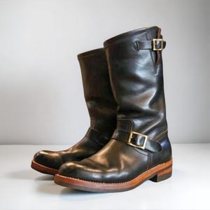 Puede incluir: Un par de botas de cuero negro para motocicleta con hebillas doradas. Las botas tienen una caña alta y una suela marrón. El cuero parece desgastado, lo que sugiere durabilidad y estilo. Las botas miden unos 30 cm de alto.