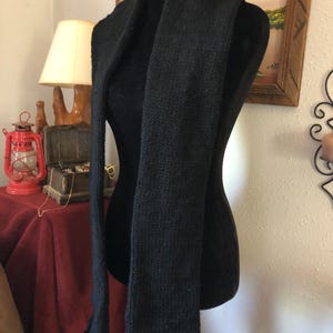 May include: A long, solid black knitted scarf with a textured, ribbed stitch pattern. The scarf is draped vertically over a black mannequin torso, featuring straight edges and a consistent dark color throughout the entire length of the garment.