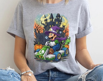 Może przedstawiać: Jasnoszara koszulka z grafiką o tematyce Halloween. Projekt przedstawia postać z kreskówki w kapeluszu czarownicy jadącą zielonym gokartem przed nawiedzonym zamkiem, dyniami i duchami. Postać ma na sobie fioletową pelerynę.