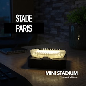 May include: A miniature stadium lamp with the text "STADE PARIS" and "MINI STADIUM". The lamp is white and sits on a black base. The stadium's interior is illuminated, creating a warm glow.