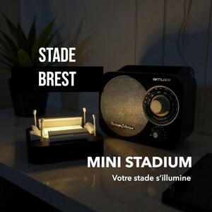 May include: A black mini stadium model with a light inside, next to a black vintage radio. The radio has a circular speaker and a dial. Text on the image reads "STADE BREST" and "MINI STADIUM".