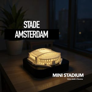 Op de afbeelding: Een verlicht miniatuurstadionmodel met de tekst "STADE AMSTERDAM" en "MINI STADIUM". Het witte stadion is van binnenuit verlicht, staat op een zwarte basis en is geplaatst op een houten oppervlak. Een plant in een pot staat op de achtergrond.