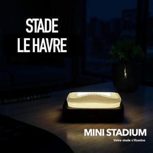 Könnte beinhalten: Ein leuchtendes Mini-Stadion-Nachtlicht mit dem Text "STADE LE HAVRE" und "MINI STADIUM" in Weiß. Das Licht hat ein Stadiondesign und steht auf einem schwarzen Sockel. Der Hintergrund ist dunkel.