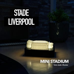 May include: A glowing miniature stadium model on a black base. The text "STADE LIVERPOOL" is above the model, with "MINI STADIUM" below. A small plant in a dark pot is in the background. The stadium is lit up.