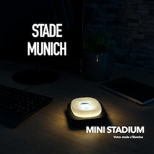 Pode incluir: Uma luz noturna em miniatura em forma de estádio com um interior brilhante, sobre uma base preta. O texto "MINI STADIUM" está abaixo da luz. As palavras "STADE MUNICH" estão em branco no canto superior esquerdo.
