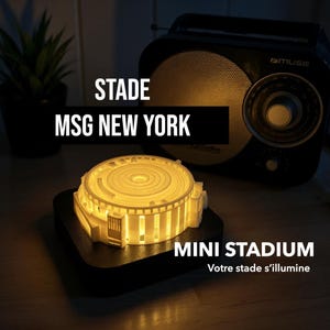 May include: A miniature stadium model with a warm, glowing interior, set on a black base. The text "STADE MSG NEW YORK" is displayed above the stadium. A vintage radio is in the background. The words "MINI STADIUM" are also visible.