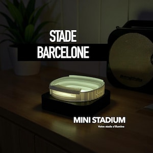 May include: A small, illuminated stadium model in white, with intricate detailing, rests on a dark base. The text "STADE BARCELONE" and "MINI STADIUM" are displayed in white. The overall theme is a miniature sports venue.