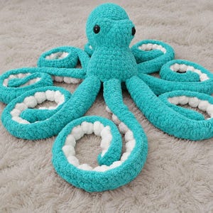 Könnte beinhalten: Ein Plüsch-Oktopus aus türkisfarbenem Garn mit weicher, flauschiger Textur. Das Wesen hat einen abgerundeten Kopf mit zwei schwarzen, kreisförmigen Augen und acht geringelten Tentakeln. Jeder Tentakel zeigt eine kontrastierende weiße Unterseite mit Noppen.