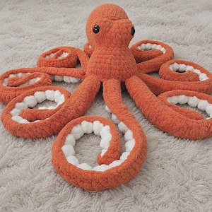 May include: A plush octopus toy crafted from orange yarn with a soft, textured finish. The creature features a rounded head with two black plastic eyes and eight curled tentacles, each displaying a white, bumpy underside made of plush material.