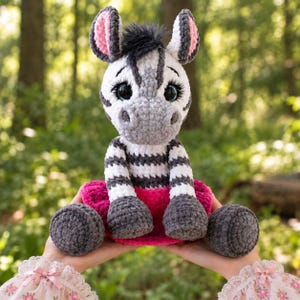 Puede incluir: Un peluche de cebra tejido a ganchillo con cuerpo de rayas blancas y grises, un mechón de pelo negro en la cabeza, ojos grandes y expresivos con pestañas largas, orejas interiores rosas y una falda texturizada de color rosa brillante en la cintura.