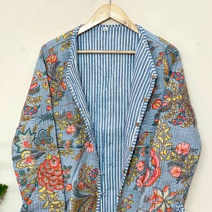 Könnte beinhalten: Hellblaue Steppjacke mit floralem Muster in Rot-, Gelb- und Grüntönen. Das Innenfutter hat blau-weiße vertikale Streifen. Die Jacke hängt an einem Holzbügel.