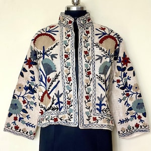 Könnte beinhalten: Cremefarbene Jacke mit aufwendiger Blumenstickerei in Blau, Rot und Beige. Die Jacke hat einen Stehkragen und Dreiviertelärmel. Die Stickerei bildet einen auffälligen Kontrast zum neutralen Hintergrund und zeigt einen Bohemian-Stil.