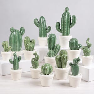 May include: A collection of various green ceramic cactus figurines, each potted in a small white container. The cacti feature different shapes, sizes, and textures, including ribbed and dotted surfaces, arranged in a group on white display blocks.