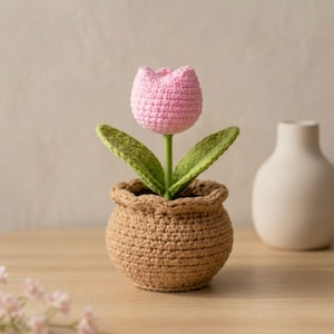 Op de afbeelding: Een gehaakte tulp in een bruine pot. De roze bloem heeft groene bladeren en een groene stengel. De pot is lichtbruin van kleur en heeft een gerimpelde rand. De achtergrond is neutraal beige.