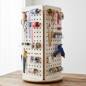 Può includere: Un organizer rotante per artigianato con finitura color crema. Presenta pannelli forati con vari materiali per l'artigianato, tra cui nastri, forbici, pennelli, barattoli di perline e una pistola per colla. L'organizer poggia su una base di legno.