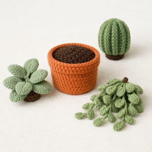 May include: Crocheted miniature plants including a potted succulent, a cactus, and a hanging plant. The plants are in shades of green and brown, with an orange pot filled with brown soil. The items are arranged on a white surface.
